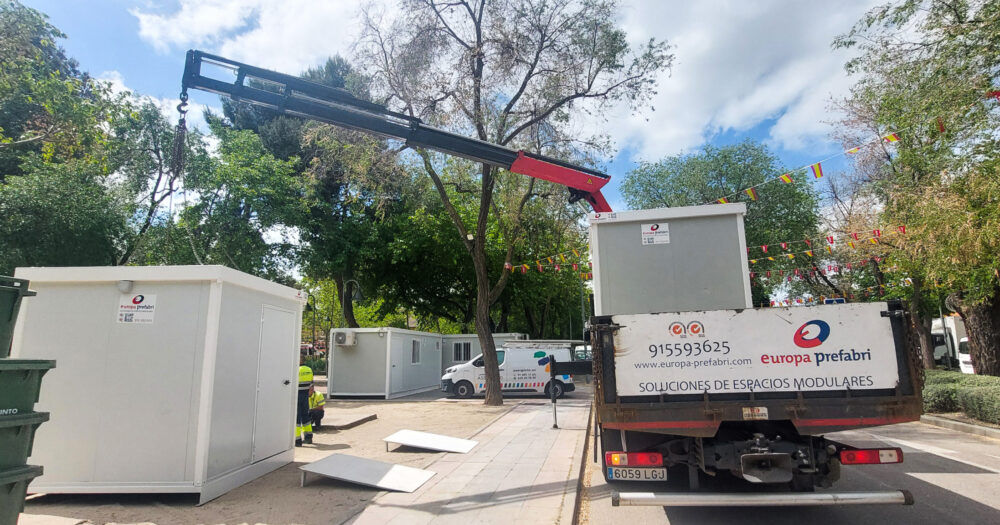 Vista frontal de dos módulos prefabricados tipo camerino instalados por Europa Prefabri para el Ayuntamiento de Pinto, con acceso independiente y acabados en blanco. En el entorno se aprecia una zona urbana destinada a actividades culturales.